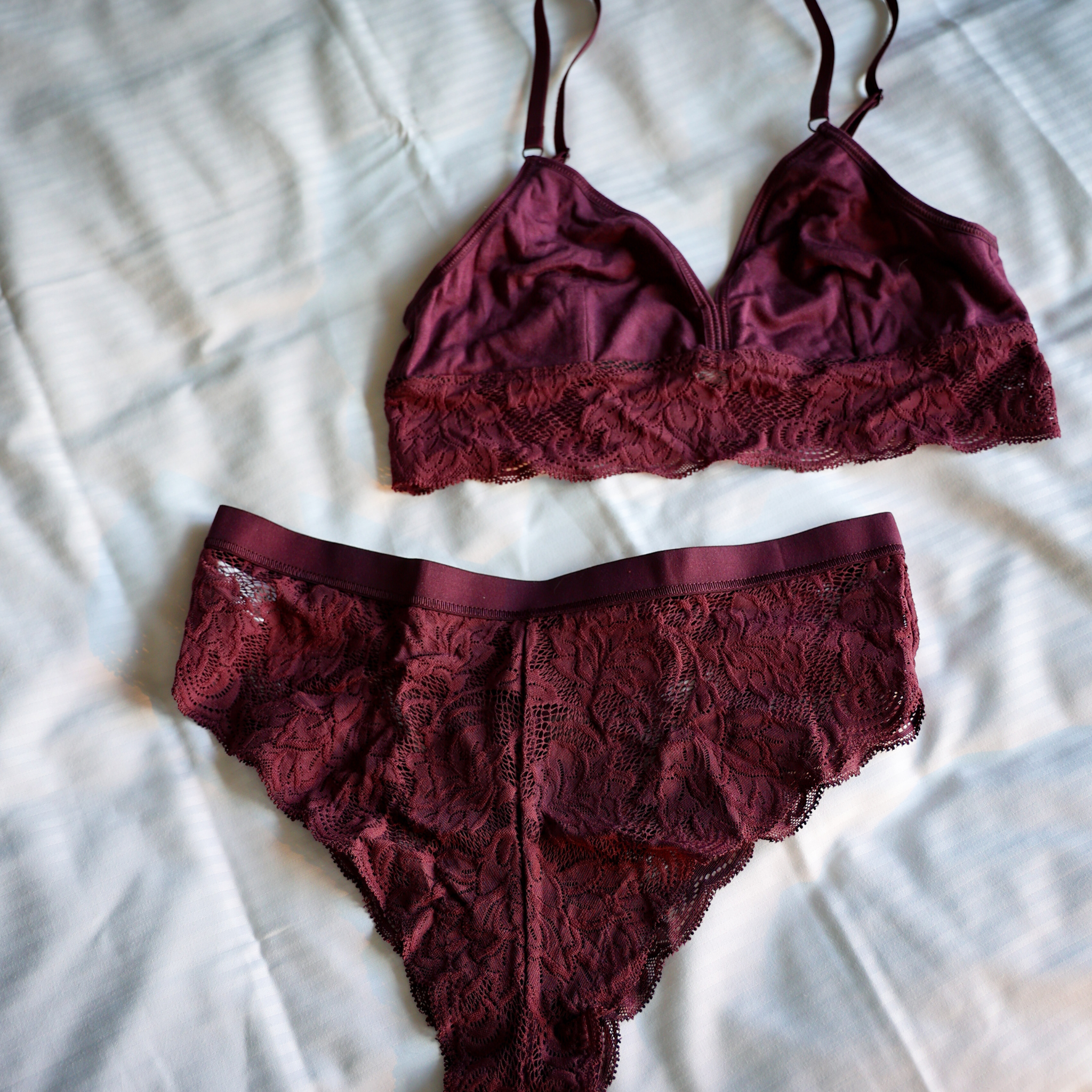 Ein passendes burgunderfarbenes Dessous-Set mit Spitzendetails liegt auf einem weißen, strukturierten Stoffhintergrund. Das Set besteht aus einem Bralette und einem Paar Höschen.
