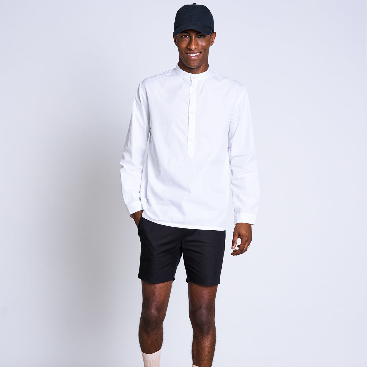 Mann in weißem Hemd und schwarzer Shorts, trägt eine schwarze Kappe und lächelt vor grauem Hintergrund.