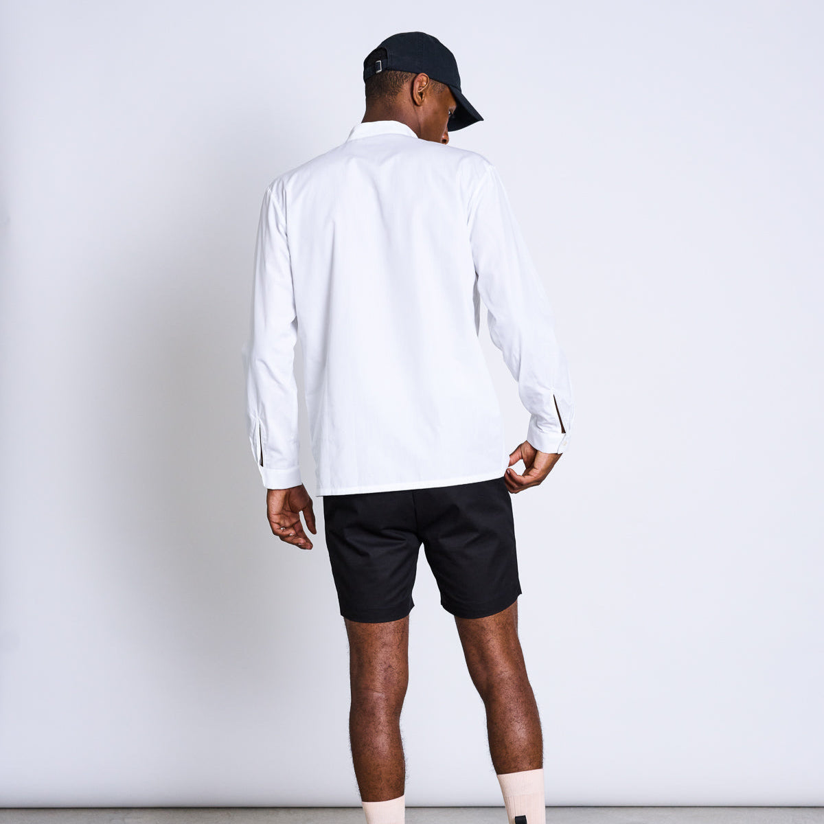 Mann von hinten in weißem Hemd, schwarzer Kappe und schwarzen Shorts, steht vor neutralem Hintergrund.