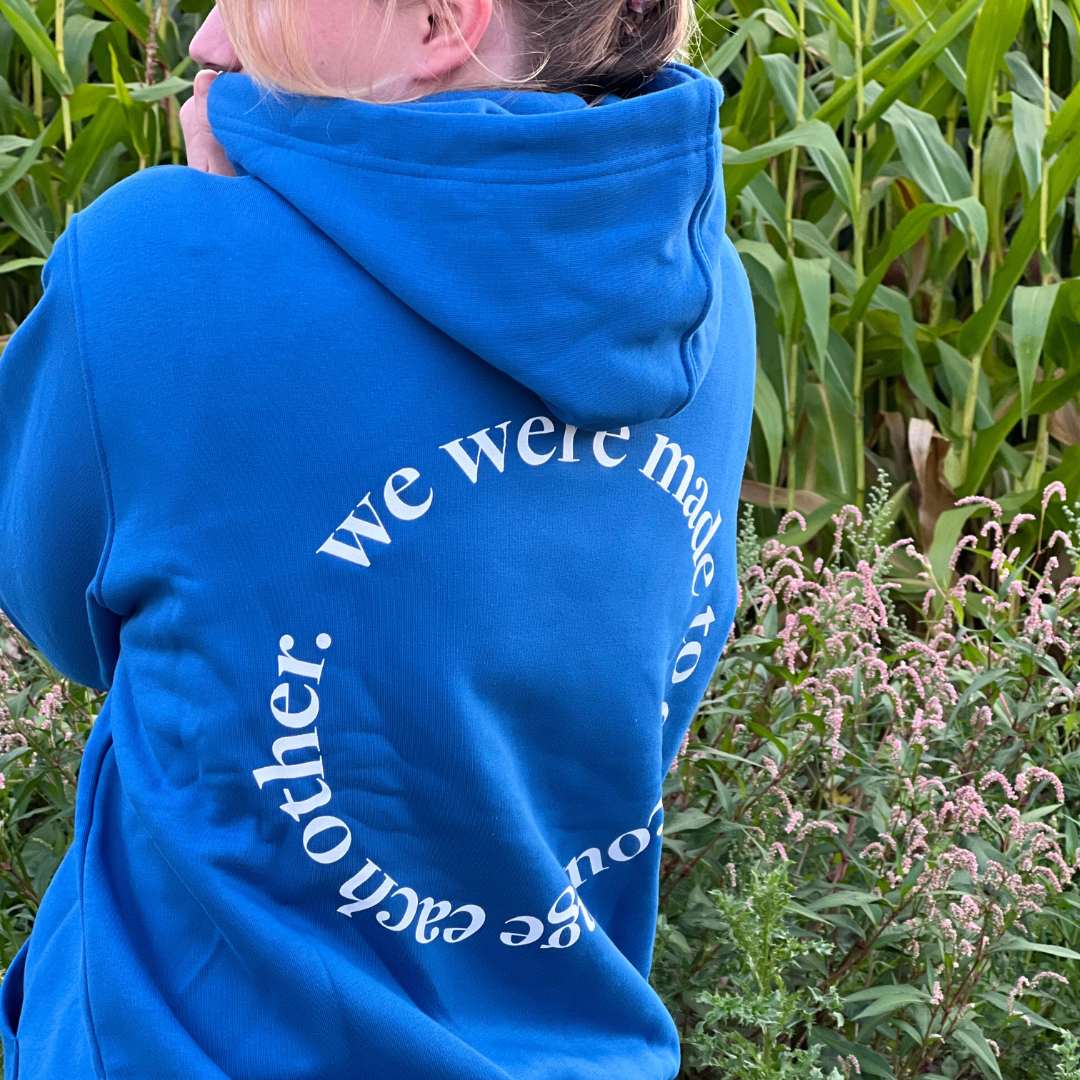 Person in a blue hoodie with text "we were made for each other" stands near tall green plants.