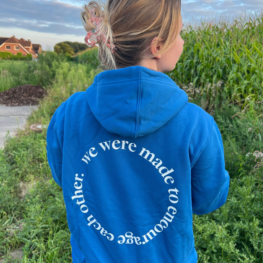 Person in blue hoodie with circular text in field, partially turned away, with houses and trees in the background.