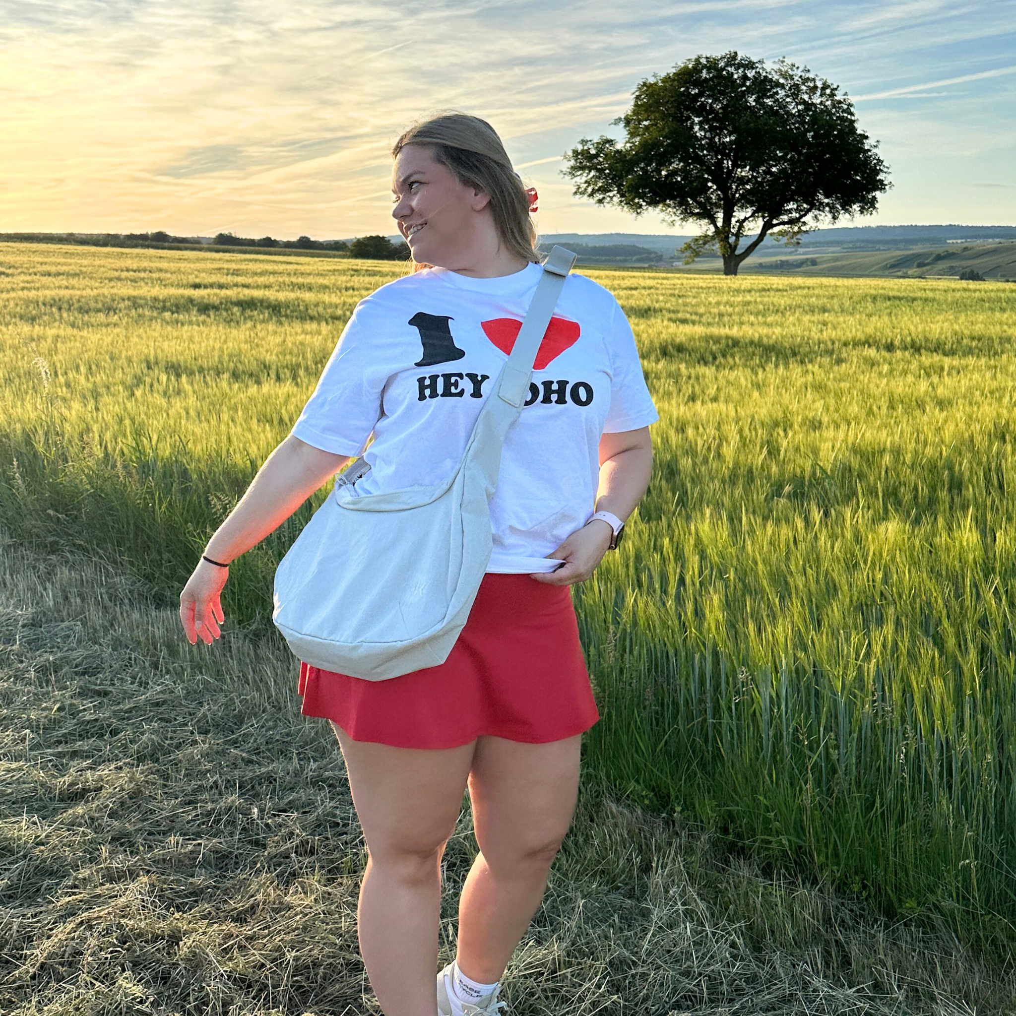 Eine Frau lächelt, während sie bei Sonnenuntergang auf einer grünen Wiese steht. Sie trägt ein weißes "I ❤️ HEY HOHO"-T-Shirt, einen roten Rock, weiße Turnschuhe und eine weiße Umhängetasche. Im Hintergrund sind ein Baum und Hügel zu sehen.