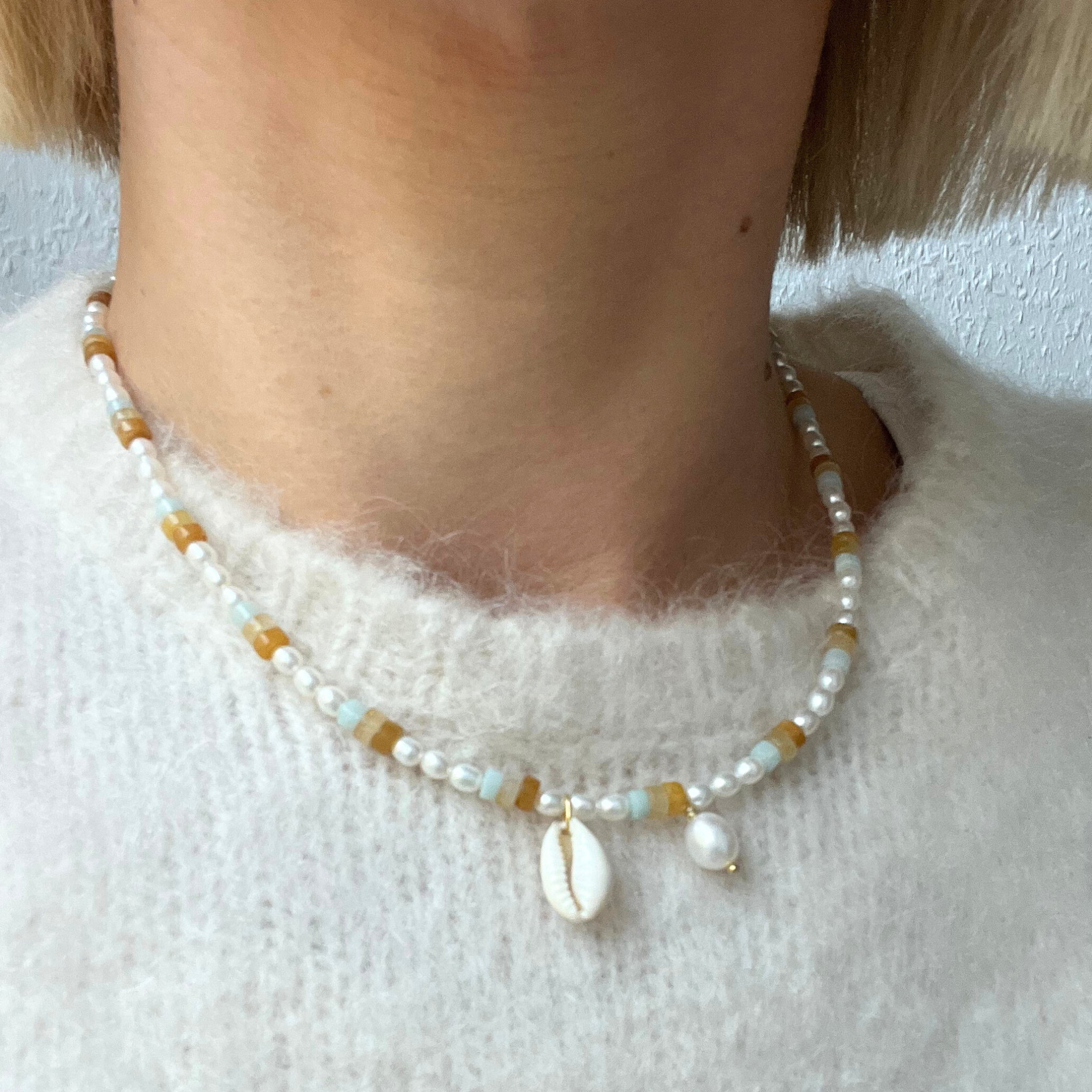 Nahaufnahme einer Halskette mit Perlen, braunen und blauen Elementen sowie einem kleinen Muschel-Anhänger, getragen über einem weißen Pullover.