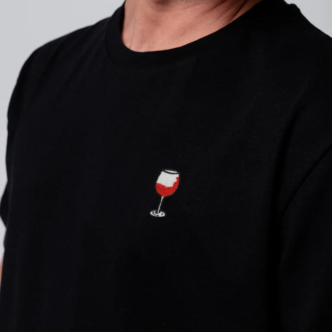 Eine Person, die ein schwarzes T-Shirt mit einem kleinen aufgestickten Rotweinglasmuster auf der Brust trägt, lächelt und schaut leicht zur Seite.