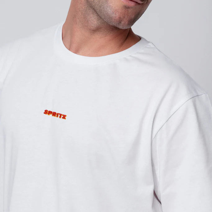 Ein Mann mit hellbraunem Haar und Schnurrbart blickt zur Seite und trägt ein schlichtes weißes T-Shirt mit dem Wort SPRITZ in kleinen orangen und roten Buchstaben auf der Brust. Der Hintergrund ist einfarbig und hell.