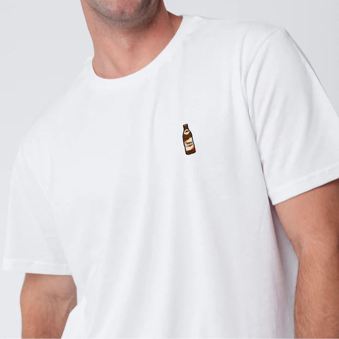 Ein lächelnder Mann mit kurzen braunen Haaren und Schnurrbart trägt ein schlichtes weißes T-Shirt mit einem kleinen aufgestickten Flaschenmotiv auf der Brust, das vor einem hellgrauen Hintergrund steht.