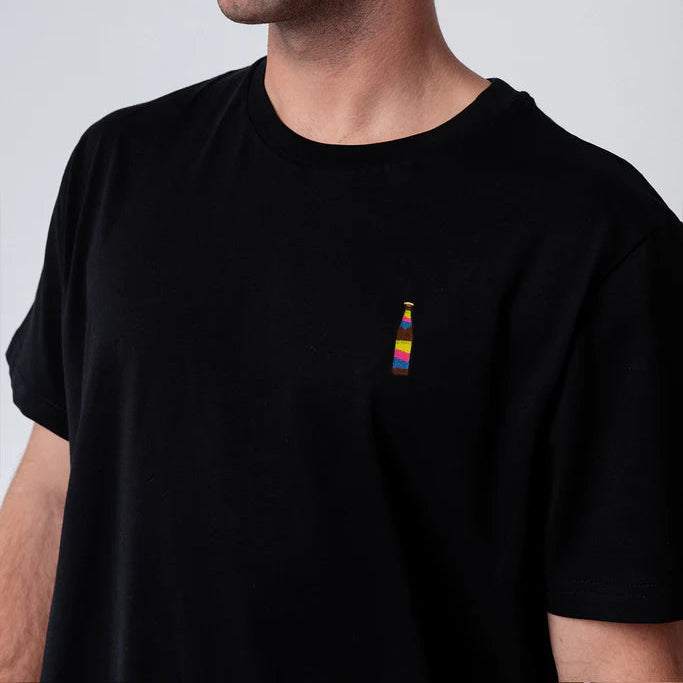 Ein Mann mit kurzen braunen Haaren, einem Schnurrbart und einem kleinen Ohrring trägt ein schwarzes T-Shirt mit einer kleinen, bunten Flaschengrafik auf der Brust. Er blickt vor einem schlichten, hellen Hintergrund zur Seite.