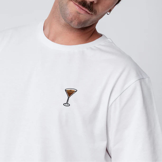 Ein Mann mit hellbraunem Haar und Schnurrbart trägt ein weißes T-Shirt mit der Abbildung eines kleinen Cocktailglases. Er neigt seinen Kopf leicht und schaut mit neutralem Blick in die Kamera.