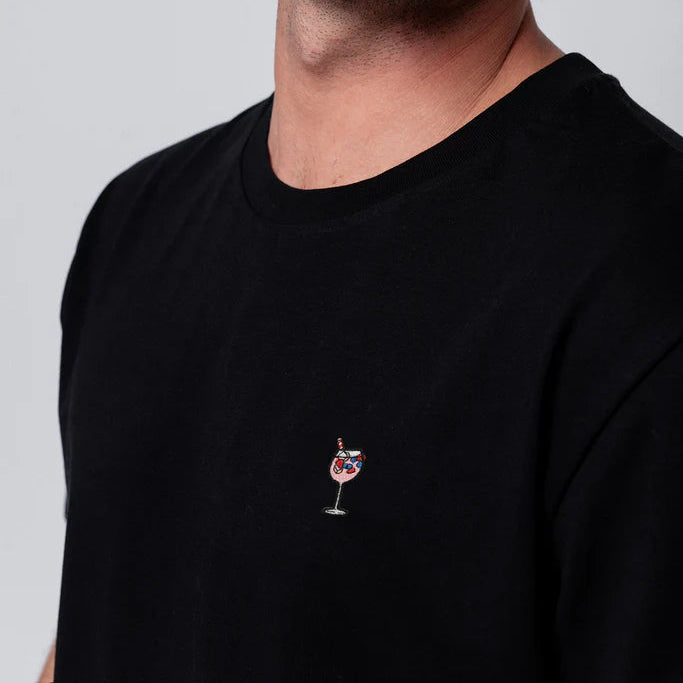 Eine Person, die ein schwarzes T-Shirt mit einem kleinen gestickten Motiv eines Cocktailglases mit Eis und einem roten Strohhalm auf der linken Seite der Brust trägt. Nur der untere Teil des Gesichts, der Hals und der obere Teil des Oberkörpers sind sichtbar.