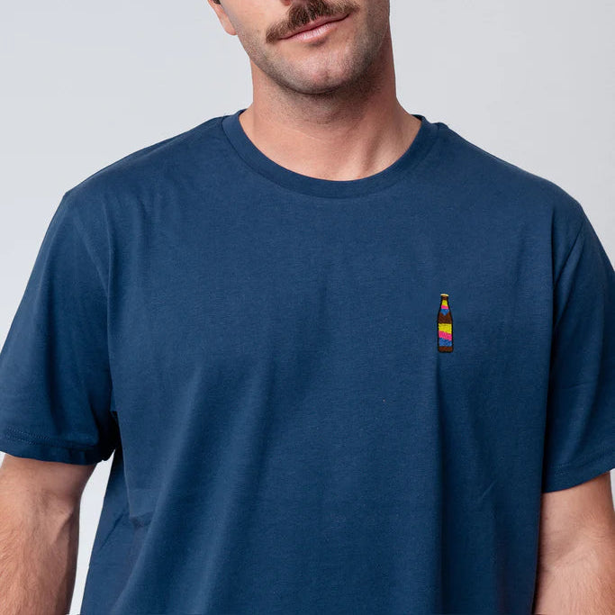 Ein Mann mit kurzen braunen Haaren und einem Schnurrbart trägt ein blaues T-Shirt mit einer kleinen, bunten Flaschengrafik auf der Vorderseite. Er hat einen neutralen Gesichtsausdruck und trägt einen silbernen Ohrring in seinem linken Ohr. Der Hintergrund ist einfarbig hellgrau.