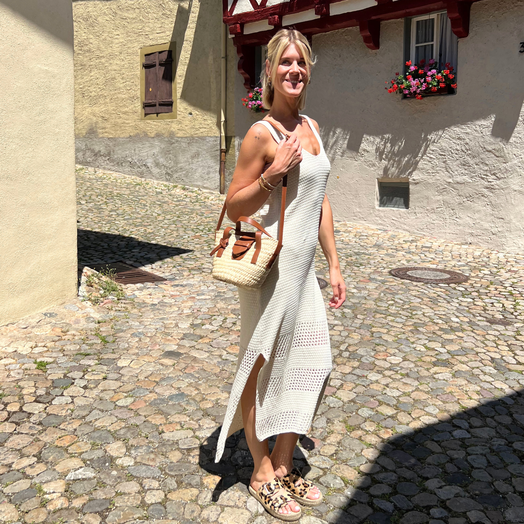 Eine Frau in einem leichten Häkelkleid und Sandalen steht auf einer sonnigen Kopfsteinpflasterstraße und hält eine gewebte Handtasche in der Hand. Sie lächelt, und bunte Blumen schmücken ein Fenster im Hintergrund.