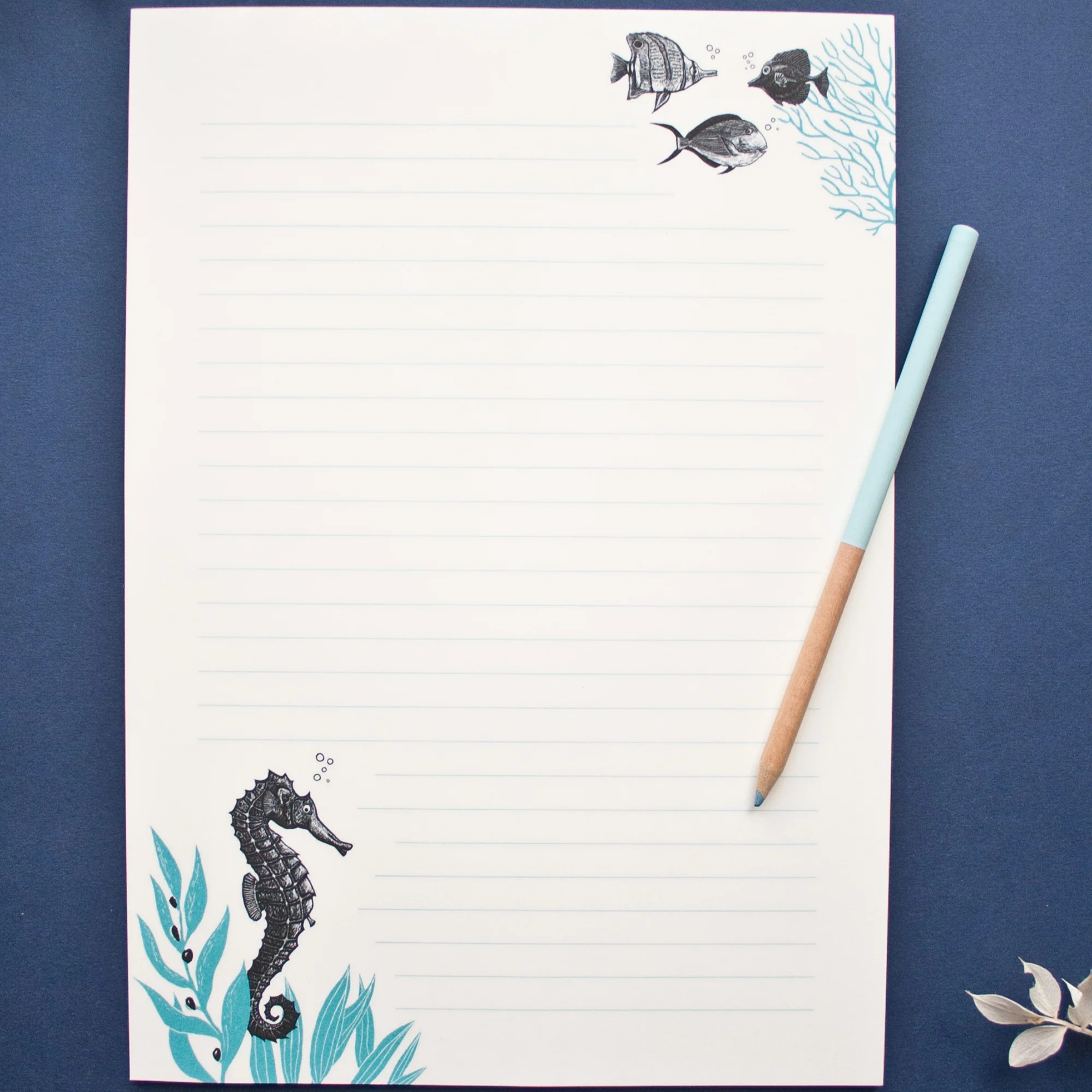 Blatt liniertes Papier mit Seepferdchen- und Fischmotiven, daneben liegt ein hellblauer Stift auf einer dunklen Oberfläche.