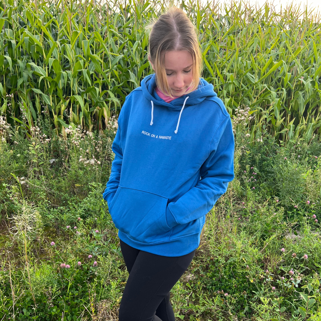 Eine Person steht in einem blauen Hoodie auf einem grünen Feld vor hohen Maispflanzen, den Blick nach unten gerichtet.