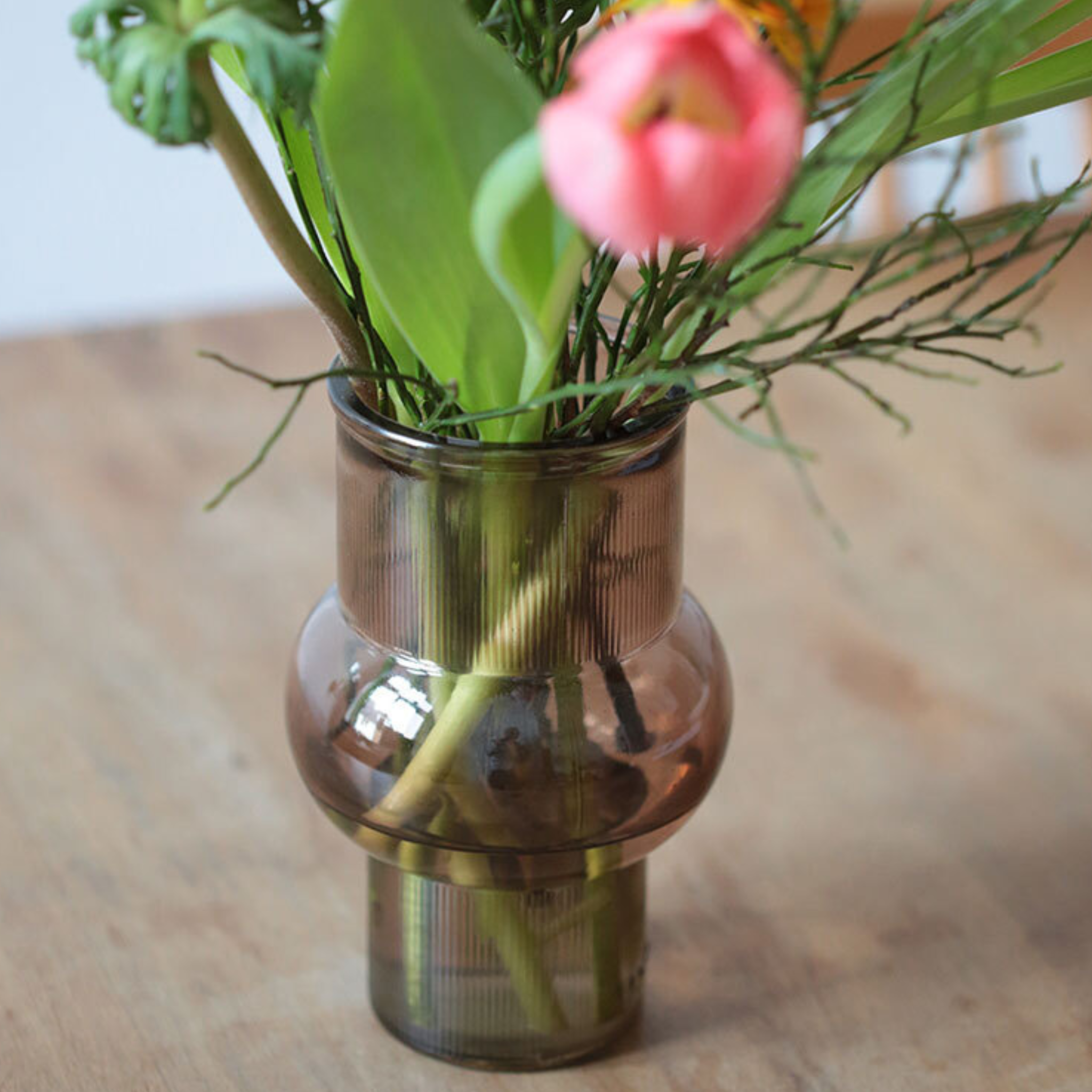 Eine braun getönte Glasvase mit abgerundeter Mitte enthält einen Blumenstrauß, darunter eine rosafarbene Tulpe und grüne Blätter, die auf einer hölzernen Unterlage stehen.