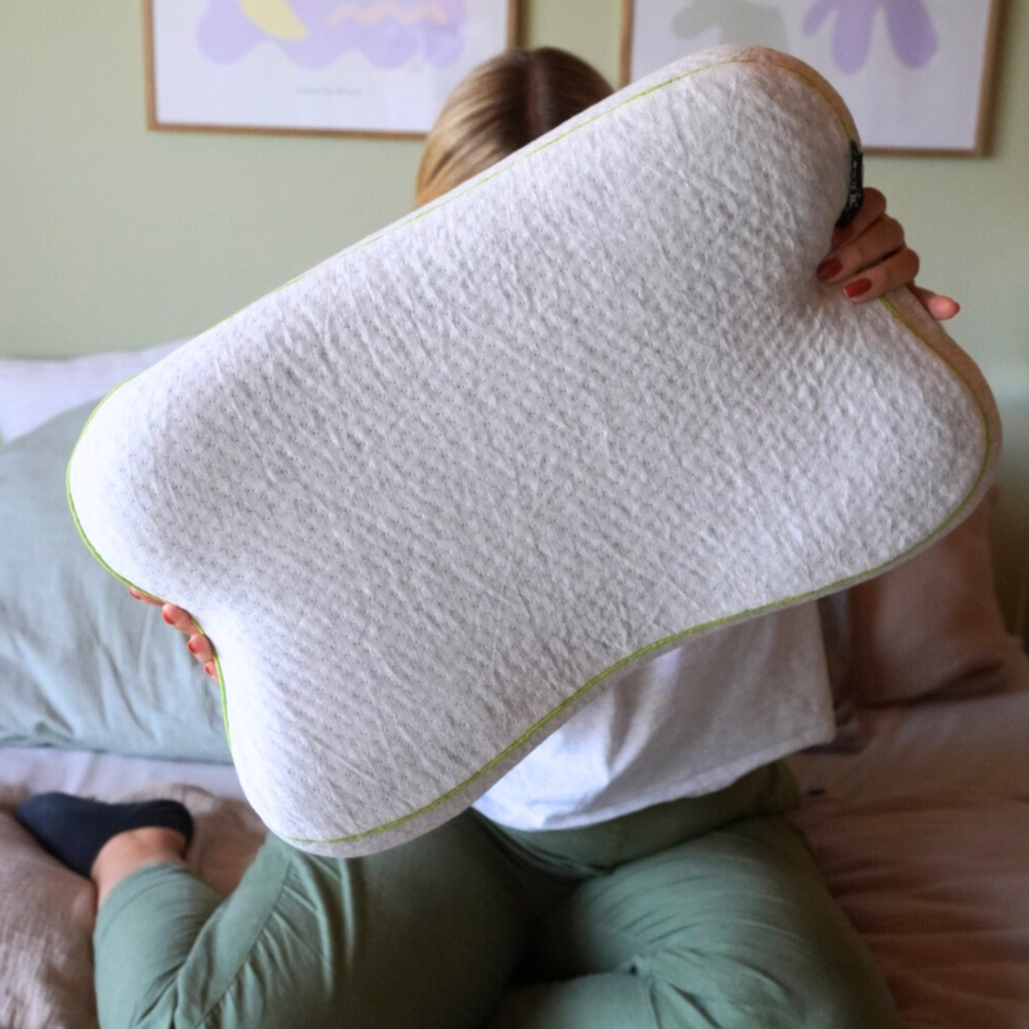 Eine Person, die auf einem Bett sitzt, hält ein großes, konturiertes Kissen hoch, das ihr Gesicht bedeckt. Die Person trägt eine grüne Hose und ein weißes Hemd. An der Wand im Hintergrund ist gerahmte Kunst zu sehen.
