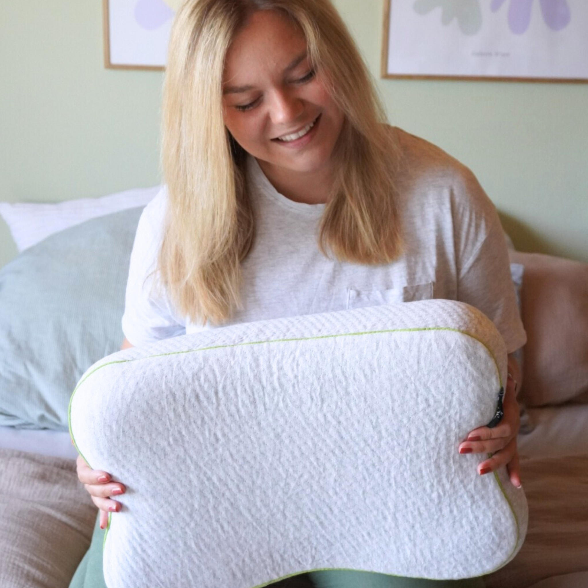 Eine Frau mit blondem Haar und weißem T-Shirt sitzt auf einem Bett und hält lächelnd ein großes, weißes, konturiertes Kissen mit grünem Rand. An der Wand hinter ihr ist ein gerahmtes Kunstwerk zu sehen.