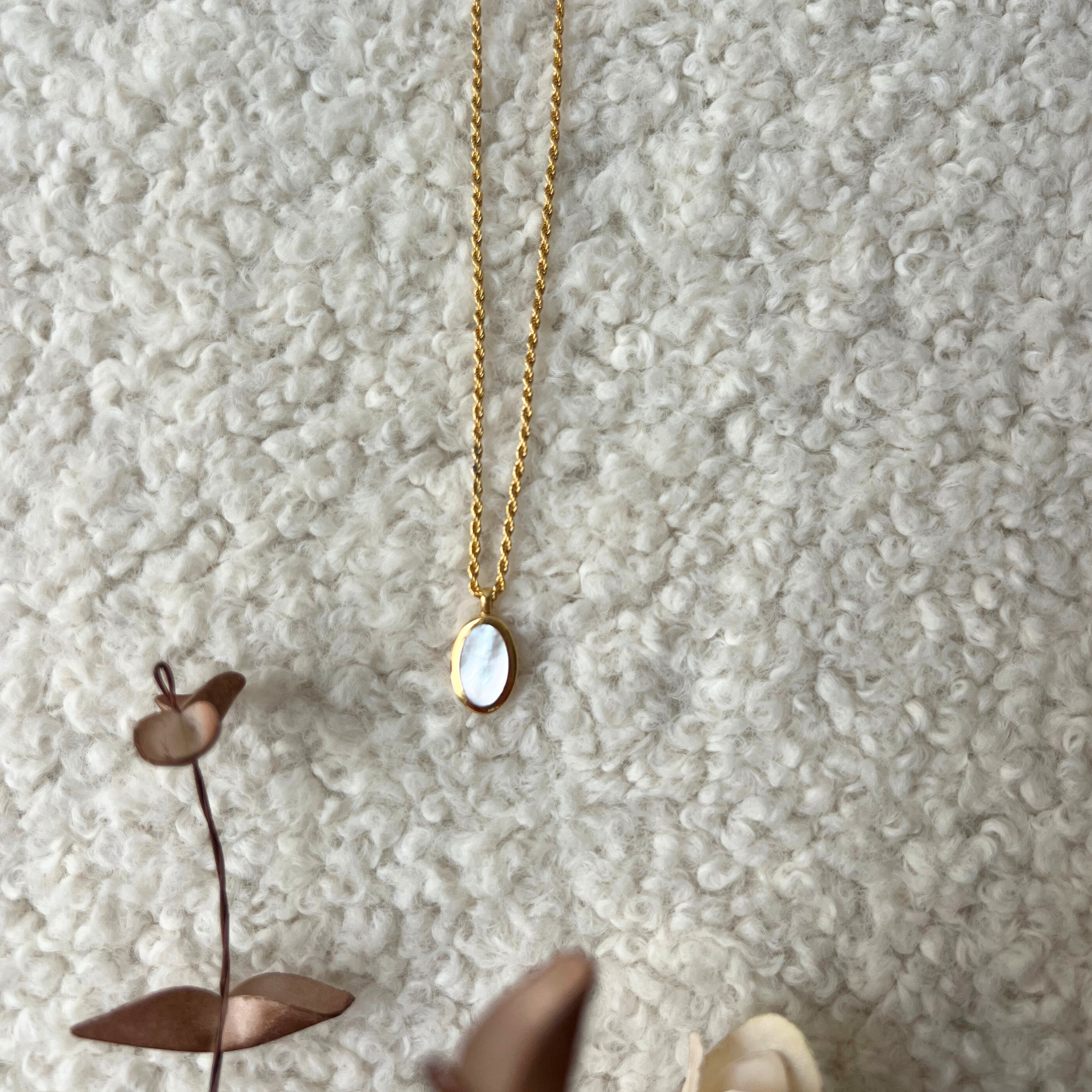 Goldene Kette mit ovalem Anhänger auf einer flauschigen cremefarbenen Oberfläche, daneben ein getrockneter Zweig.