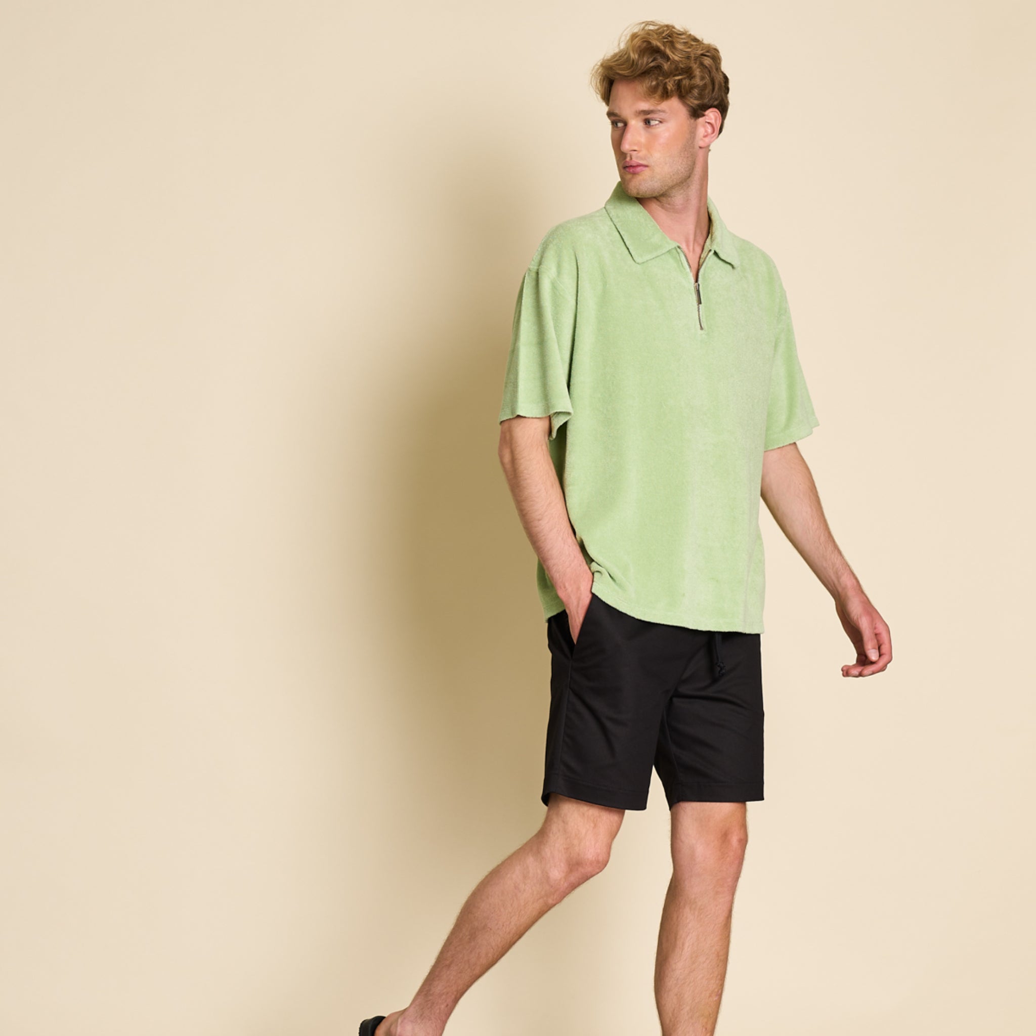 A man in a light green shirt and black shorts walks against a beige background.