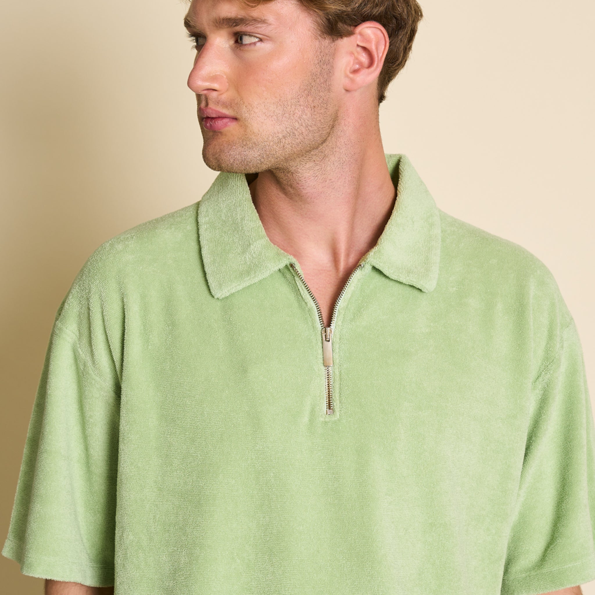 Mann in einem hellgrünen Poloshirt mit Reißverschluss und weitem Kragen, vor beigem Hintergrund, schaut zur Seite.