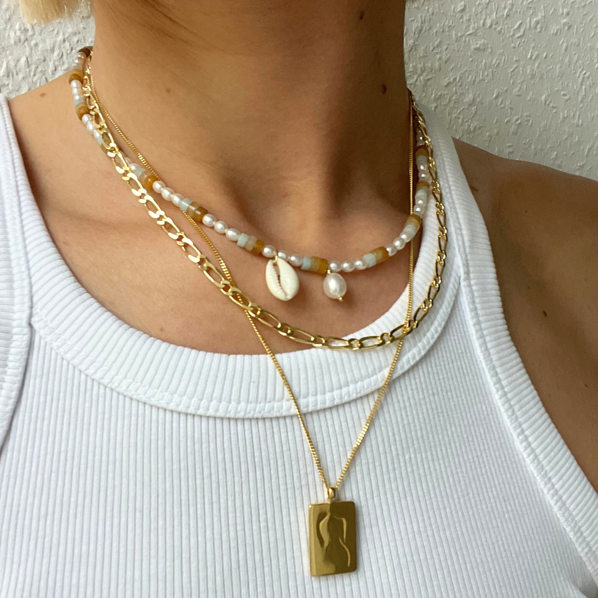 Frau trägt drei Halsketten: eine mit Perlen und Muscheln, eine goldene Kette und eine mit rechteckigem Anhänger, auf weißem Top.