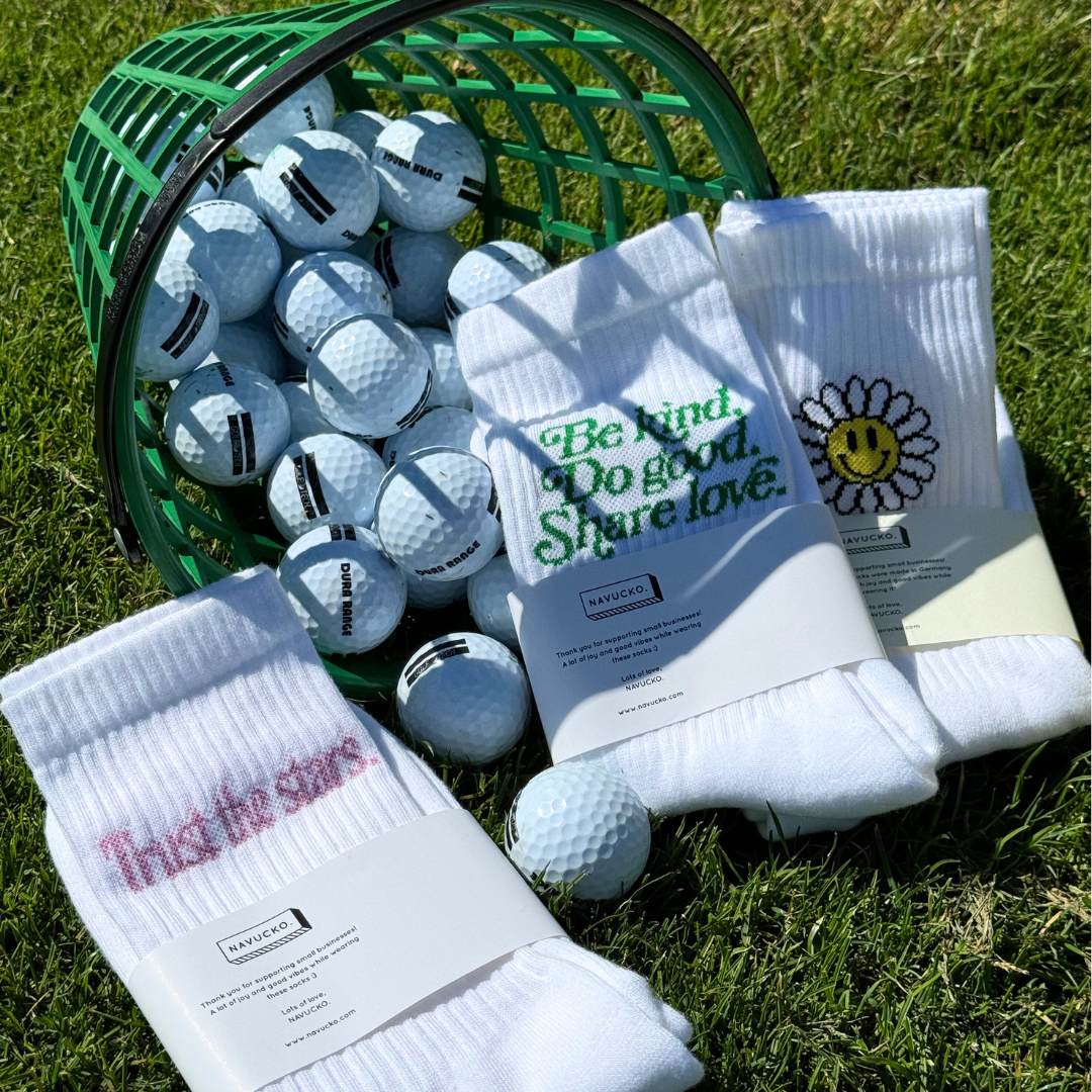 Ein grüner Korb mit Golfbällen neben drei Paar weißen Socken mit positiven Botschaften und Designs auf Gras.