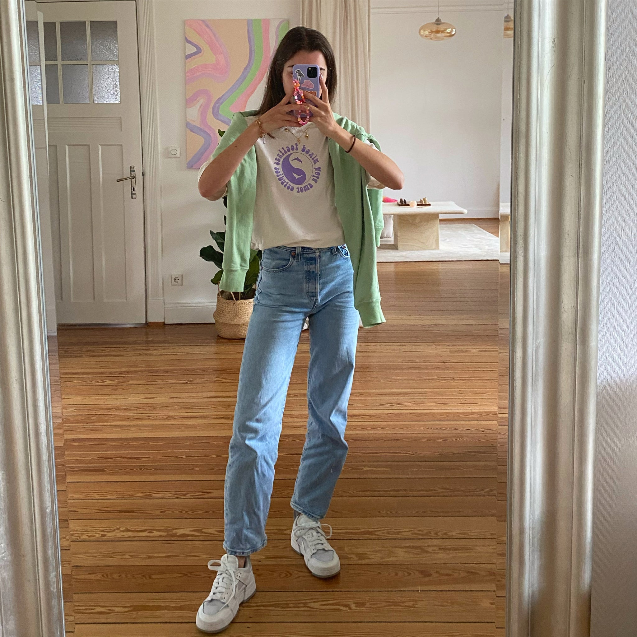 Frau macht ein Spiegel-Selfie in einem Raum mit hellem Holzboden, trägt Jeans, ein T-Shirt und eine grüne Jacke.