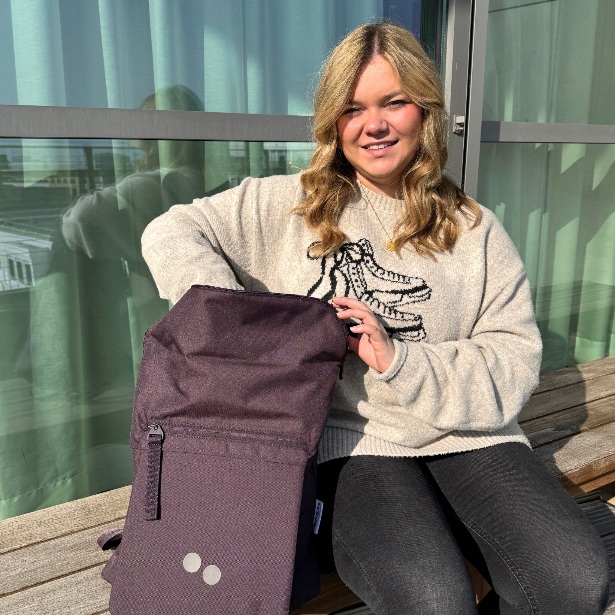 Eine Frau sitzt auf einer Bank mit einem lila Rucksack auf dem Schoß und lächelt in die Kamera.