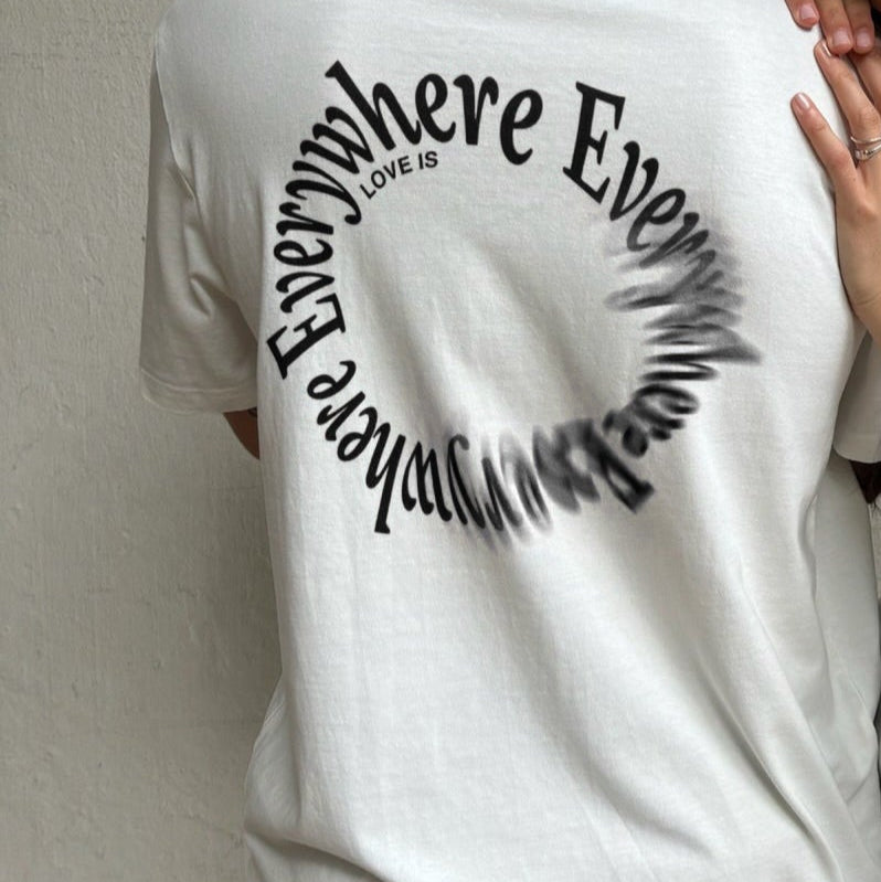 Person trägt ein weißes T-Shirt mit einem kreisförmigen Text, der "LOVE IS EVERYWHERE" in schwarzer Schrift zeigt.
