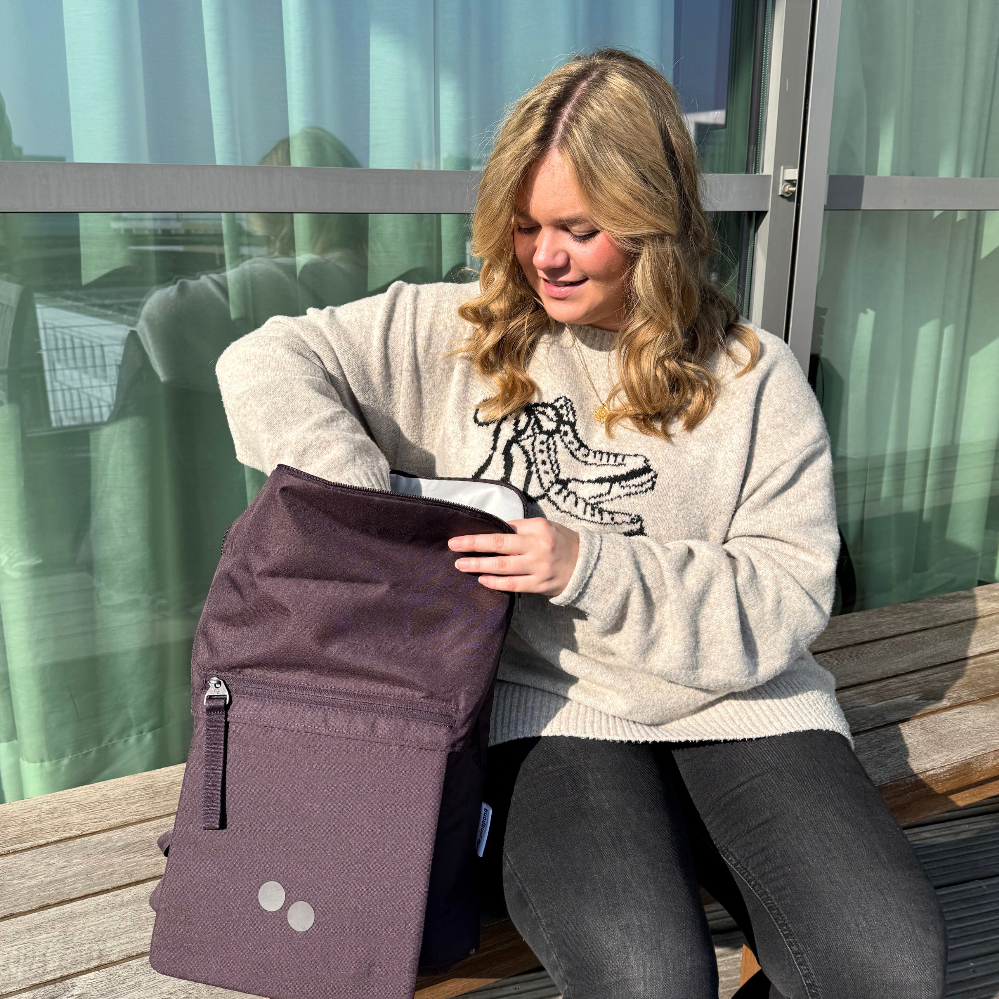 Eine Frau sitzt auf einer Holzbank und schaut in ihren lilafarbenen Rucksack, der auf ihrem Schoß liegt.