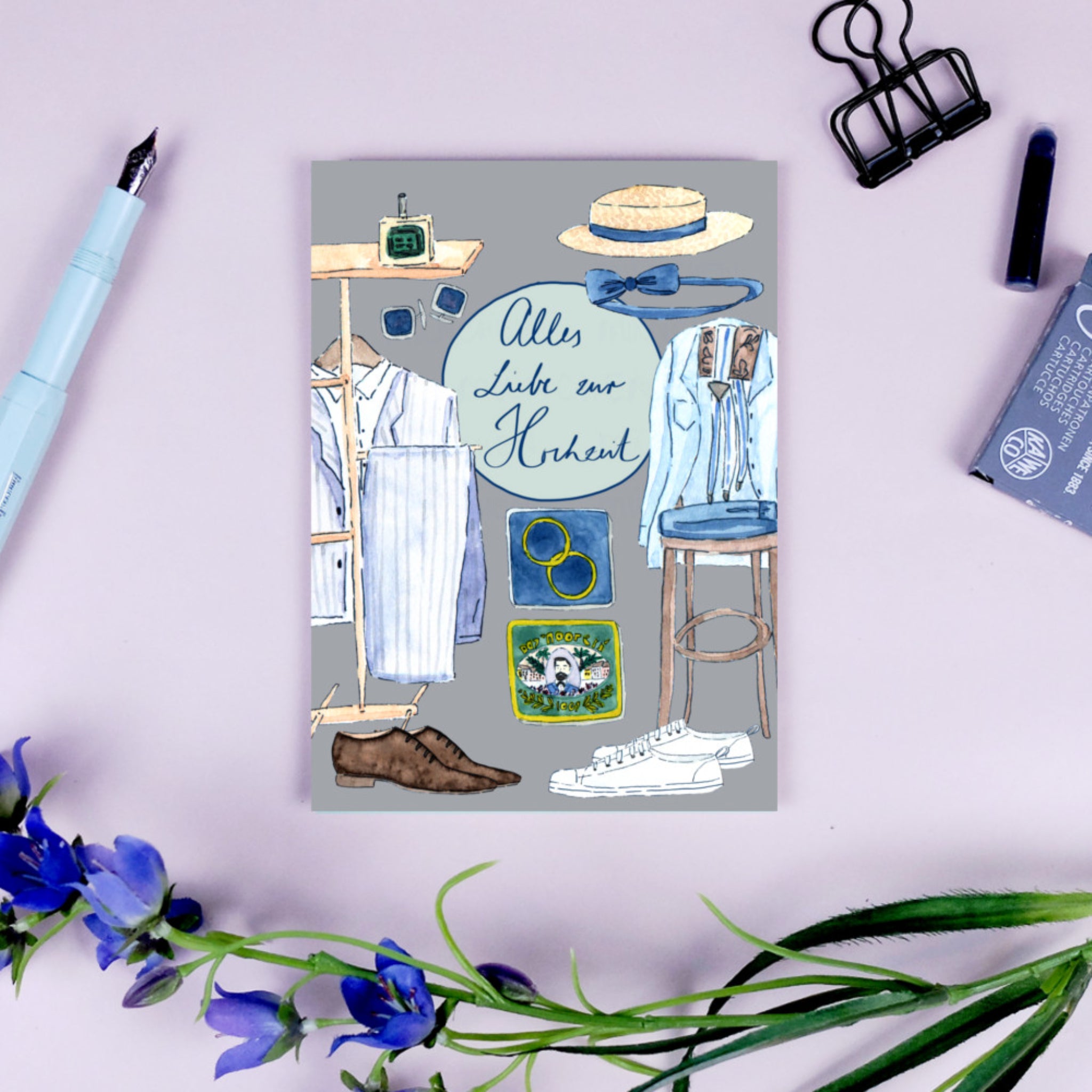Hochzeitskarte mit illustrierten Kleidungsstücken und Accessoires, Text "Alles Liebe zur Hochzeit", umgeben von Büroartikeln.