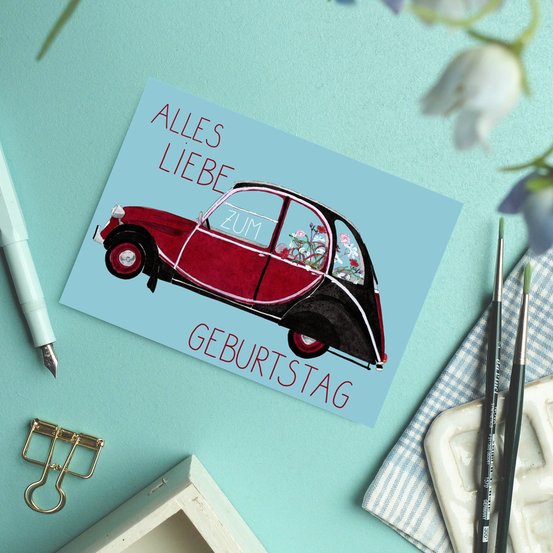 Eine Postkarte auf einem blaugrünen Schreibtisch zeigt ein illustriertes rotes Auto mit Blumen darin. Der deutsche Text lautet "Alles Liebe zum Geburtstag". In der Umgebung befinden sich ein Stift, Pinsel, eine Büroklammer und Blumen.