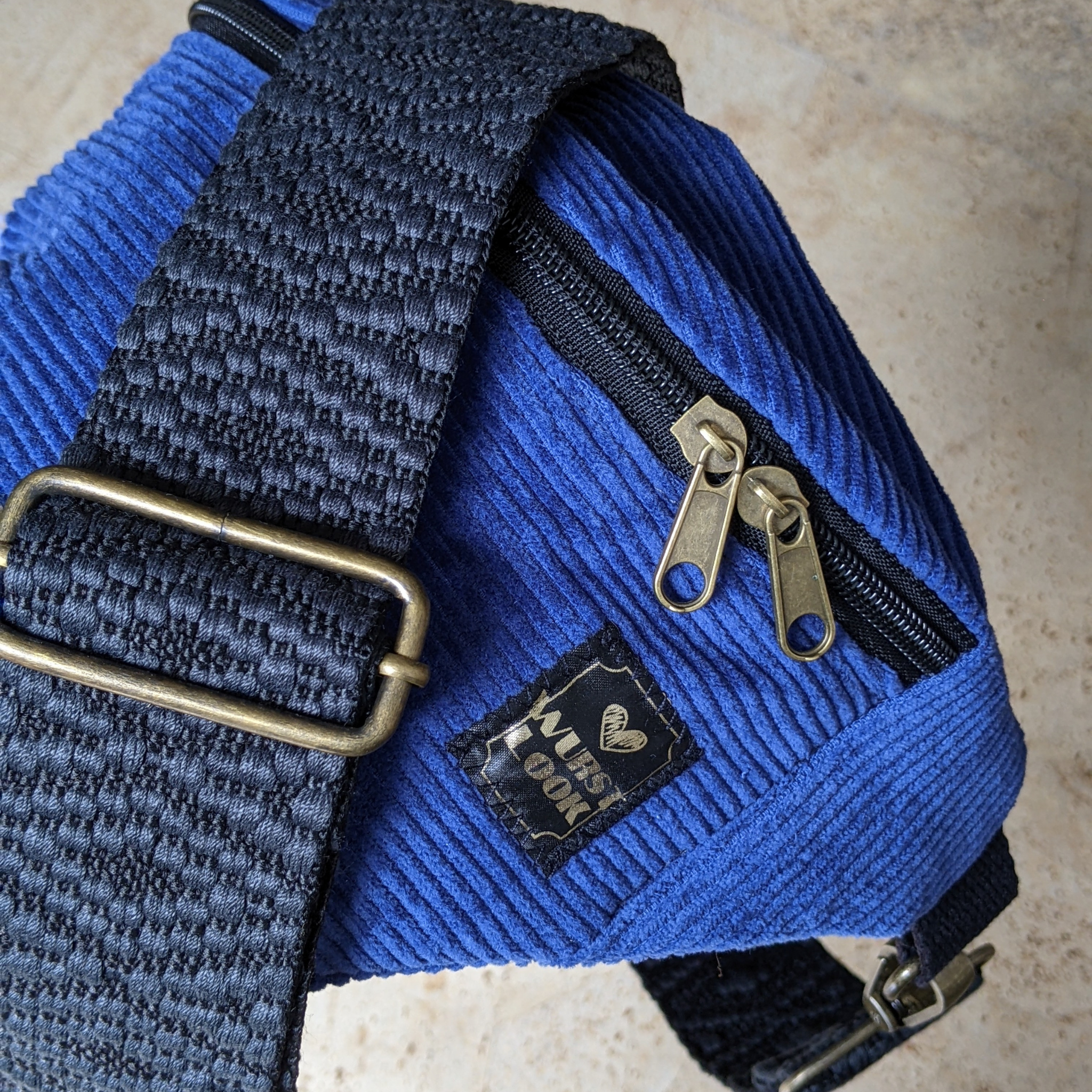 Nahaufnahme einer blauen Cordtasche mit schwarzen gewebten Riemen, Messingschnallen und Reißverschlüssen. Ein kleines schwarzes Etikett mit einem Herz und der Aufschrift LOVE YOUR PLANET ist auf die Tasche genäht.