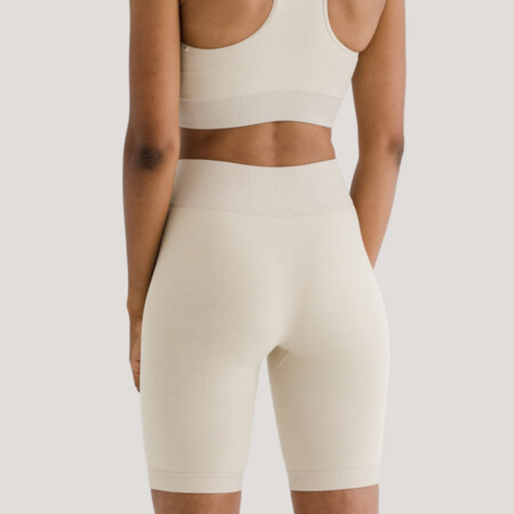 Eine Person trägt ein beigefarbenes Sportoberteil und kurze Radlerhosen, von hinten gesehen, vor einem grauen Hintergrund.