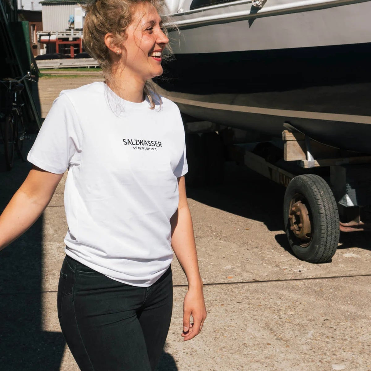 Eine Frau mit lockigem blondem Haar, die ein weißes SALZWASSER-T-Shirt und eine dunkle Hose trägt, lächelt, während sie an einem sonnigen Tag im Freien neben einem angedockten Segelboot steht.