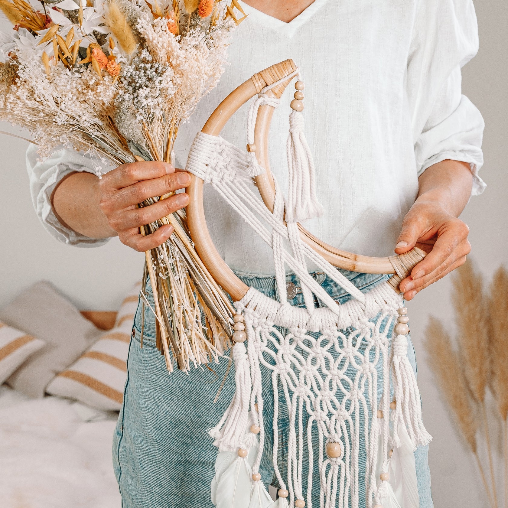 Eine Person in weißem Hemd und blauer Hose hält in der einen Hand einen Strauß getrockneter Blumen und in der anderen einen Makramee-Wandbehang mit Holzreifen und Perlen. Der Hintergrund zeigt einen gemütlichen, neutral getönten Raum.