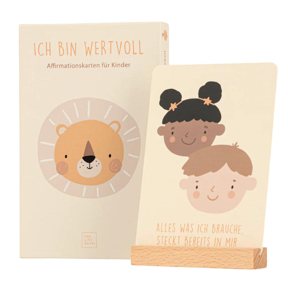 Eine Schachtel mit der Aufschrift Ich bin wertvoll mit einem Cartoon-Löwen und einer Karte auf einem Holzständer, auf der die Gesichter von zwei Kindern abgebildet sind und der Text Alles was ich brauche, steckt bereits in mir.