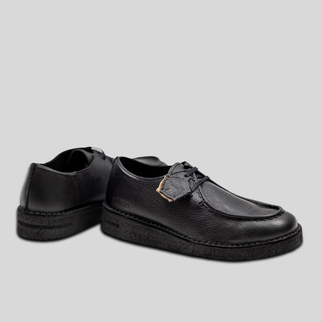 Ein Paar schwarze Lederschnürschuhe mit strukturierten Sohlen, dargestellt auf einem schlichten hellgrauen Hintergrund. Ein Schuh zeigt nach vorne, der andere ist nach hinten geneigt.