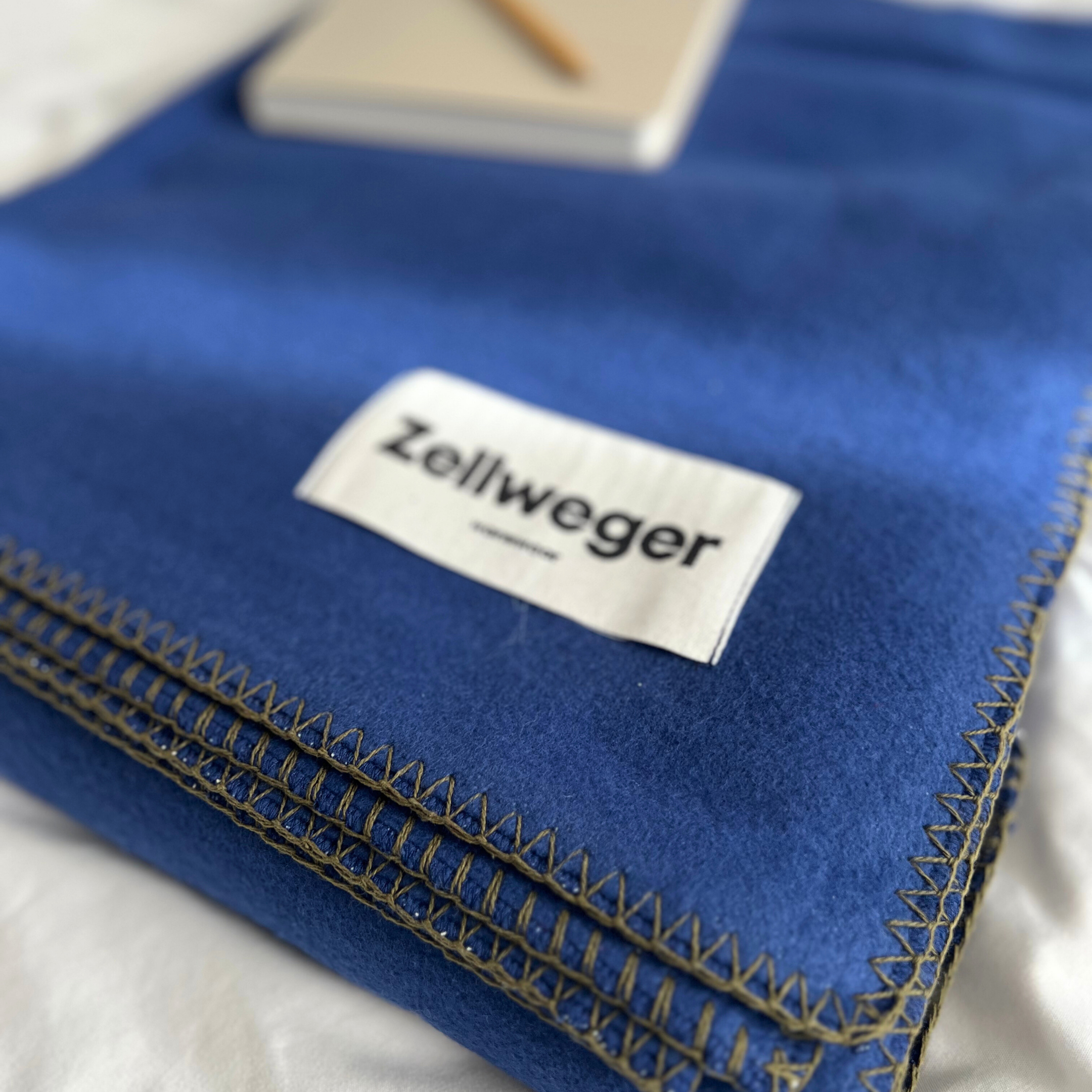 Eine blaue Decke mit einem weißen Etikett "Zellweger", gestapelt auf einem weißen Bett. Im Hintergrund liegt ein Notizbuch.