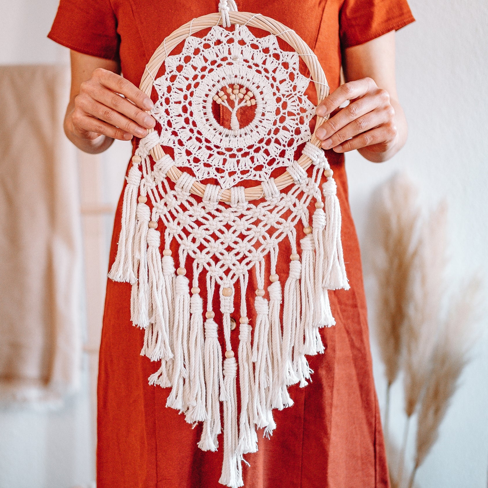 Eine Person in einem rostfarbenen Kleid hält einen handgefertigten Makramee-Wandbehang mit komplizierten Mustern, Fransen und Quasten in einem sanft beleuchteten Raum.