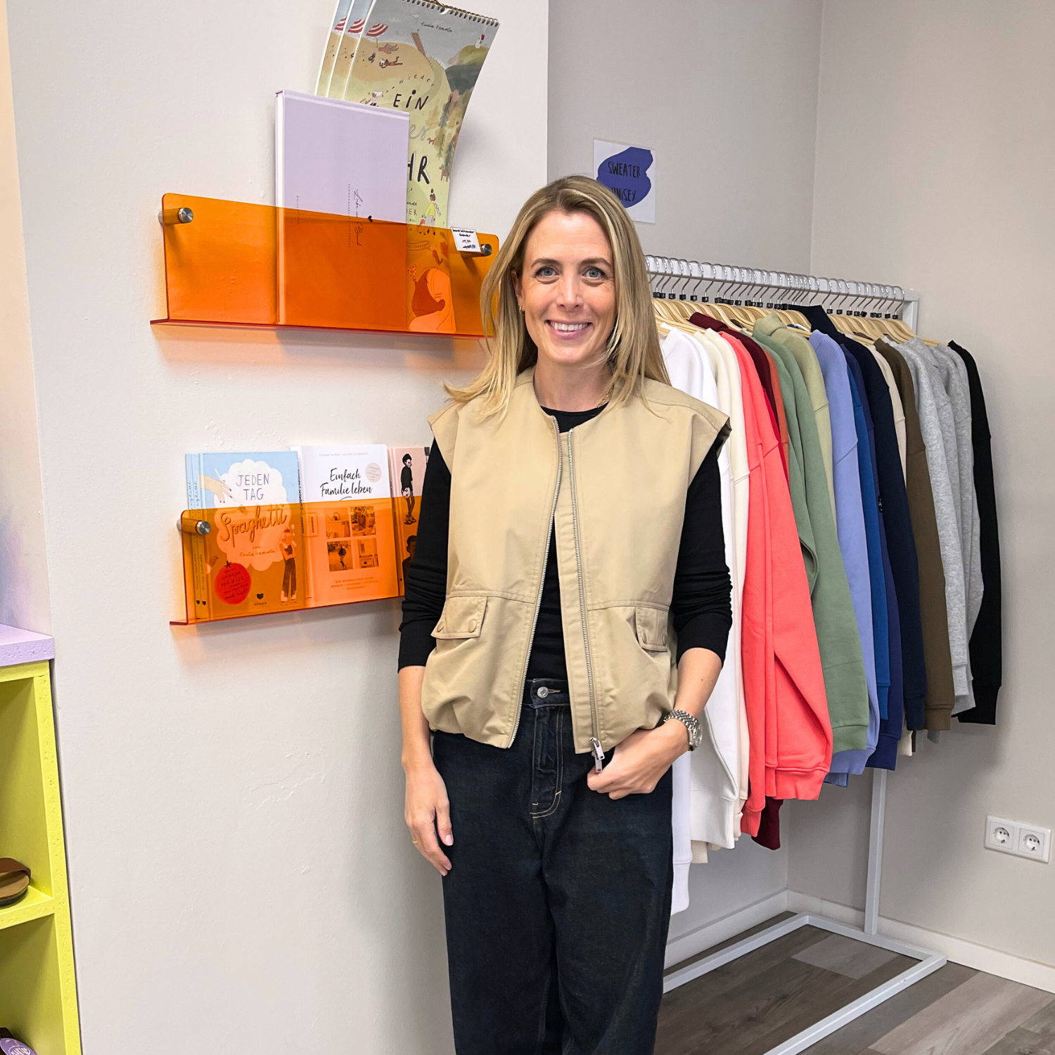Frau in beiger Weste und schwarzem Shirt steht vor Bücherregal und Kleiderständer mit bunter Kleidung.