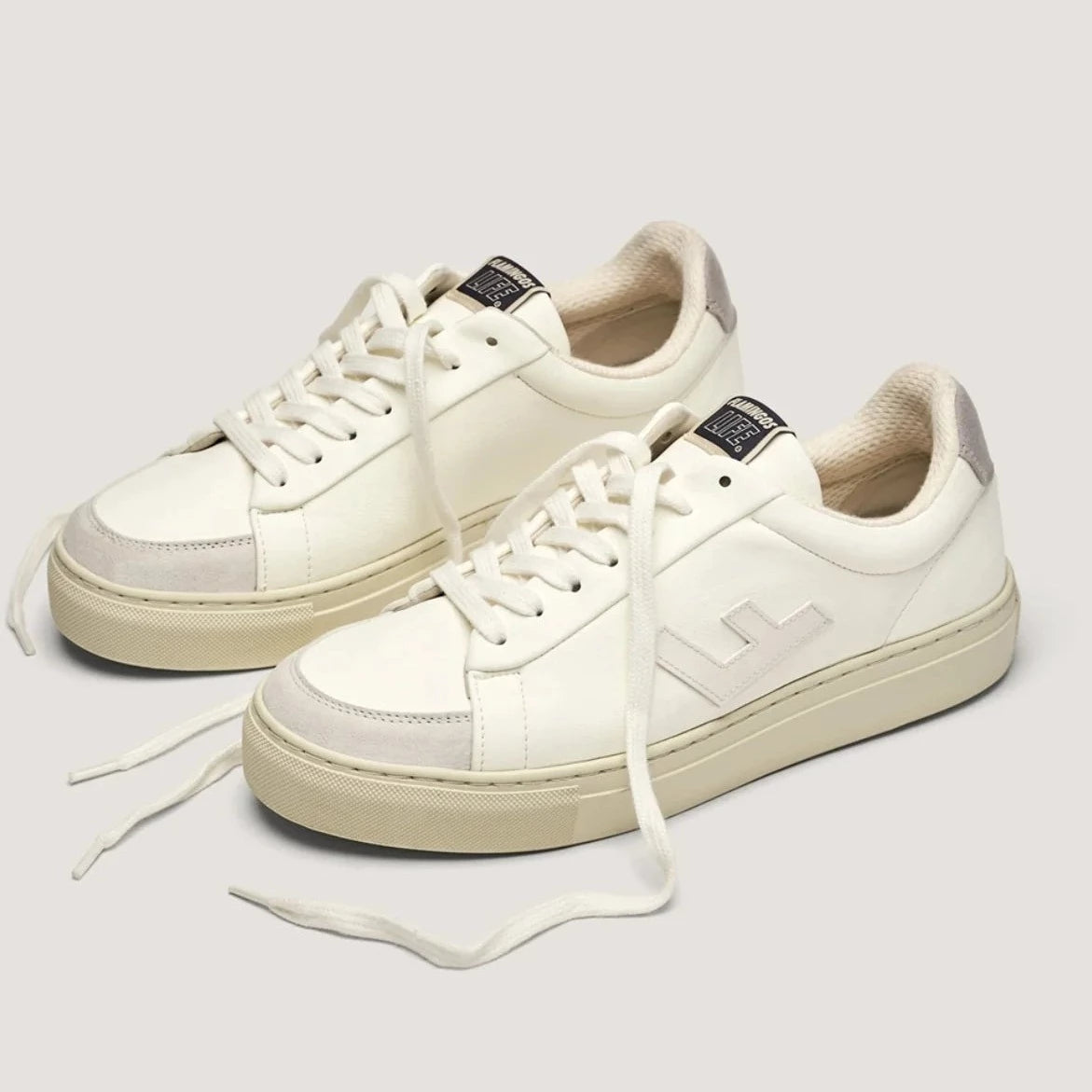 Weiße Sneaker mit Schnürsenkeln, dezentem Logo und beigem Sohlenrand vor grauem Hintergrund.