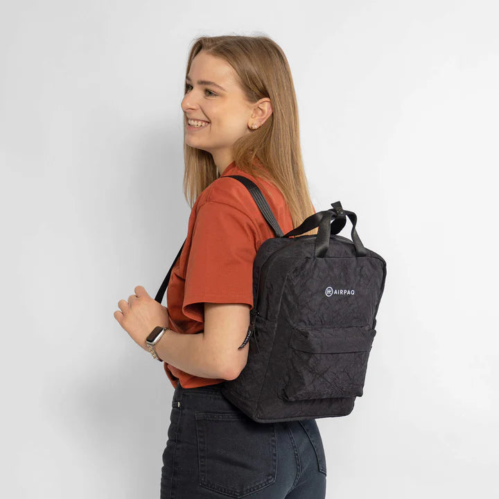 Eine lächelnde Frau mit langen Haaren trägt ein rotes T-Shirt und dunkle Jeans. Sie trägt einen schwarzen AIRPAQ-Rucksack auf dem Rücken und steht vor einem schlichten weißen Hintergrund.