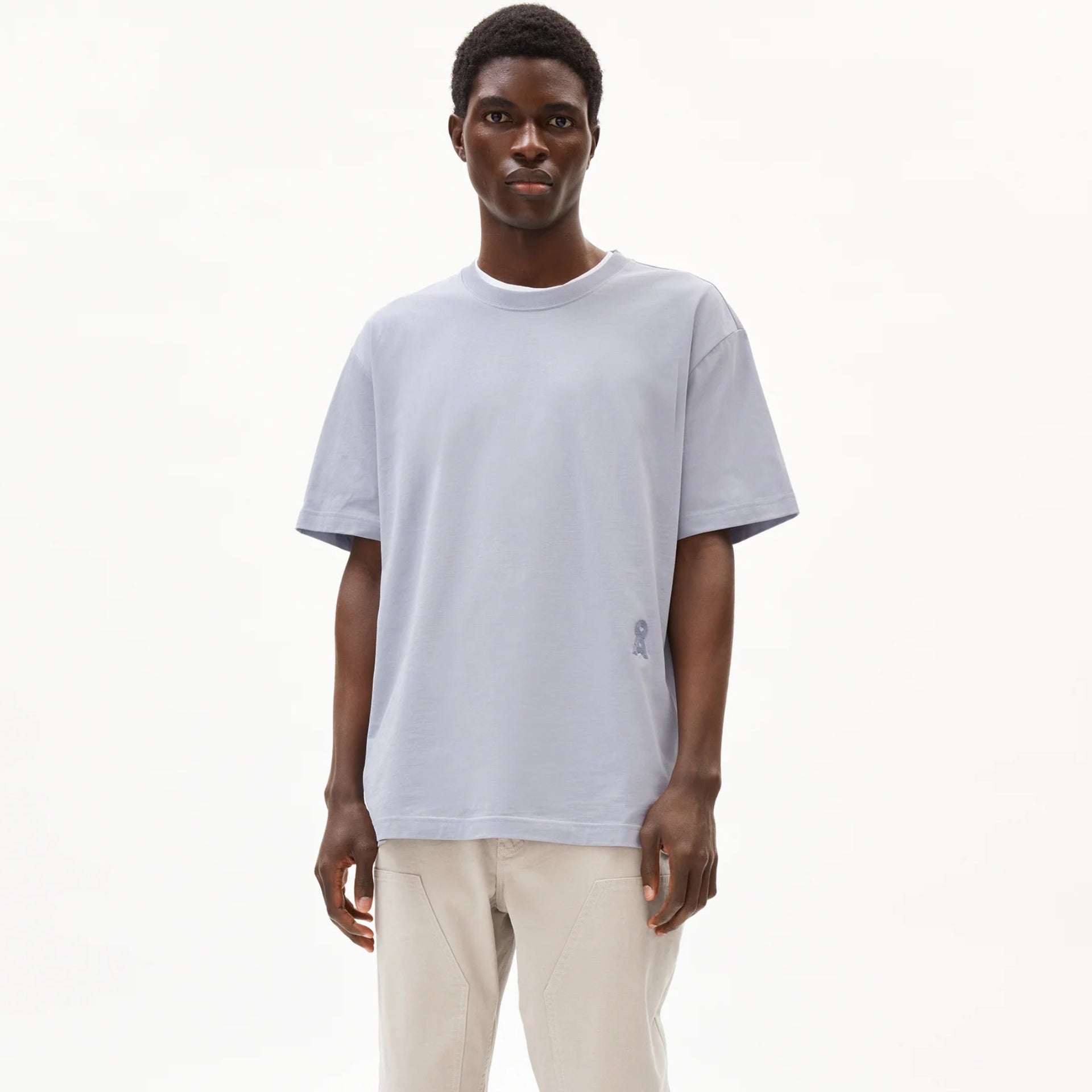 Eine Person steht vor einem schlichten weißen Hintergrund und trägt ein hellgraues, kurzärmeliges T-Shirt, eine beigefarbene Hose und graue Sportschuhe. Sie hat kurze Haare und einen neutralen Gesichtsausdruck.