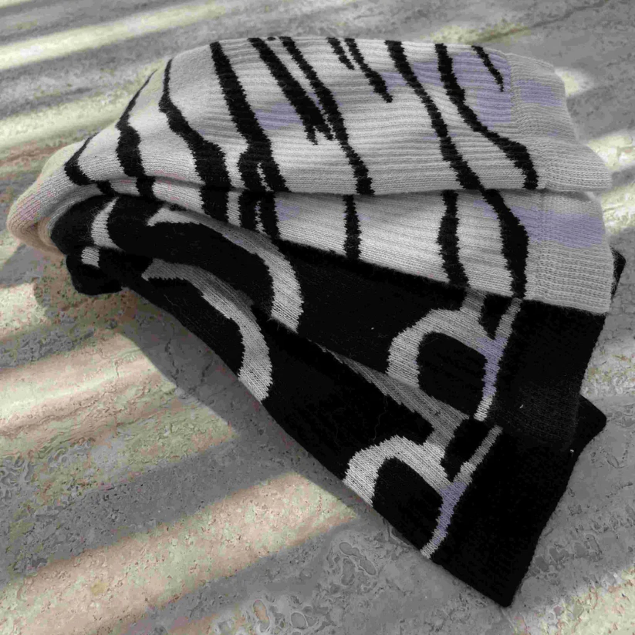 Ein gefalteter Strickschal mit abstrakten schwarzen und weißen Mustern liegt auf einer strukturierten Oberfläche, die teilweise von gestreiftem Sonnenlicht beleuchtet wird.