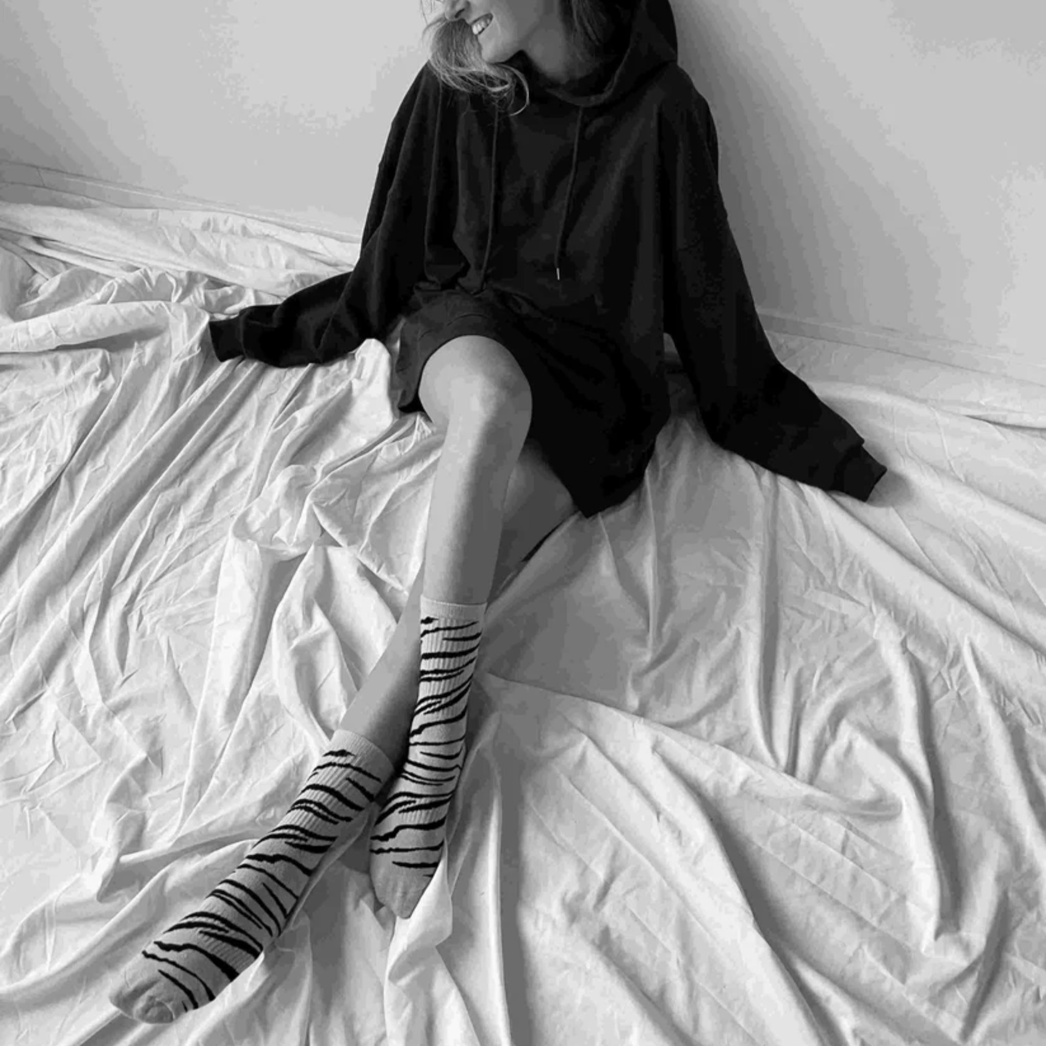 Eine Person sitzt auf einem Bett, trägt einen Kapuzenpullover und gestreifte Socken und lächelt mit gekreuzten Beinen. Das Bild ist in schwarz-weiß gehalten.