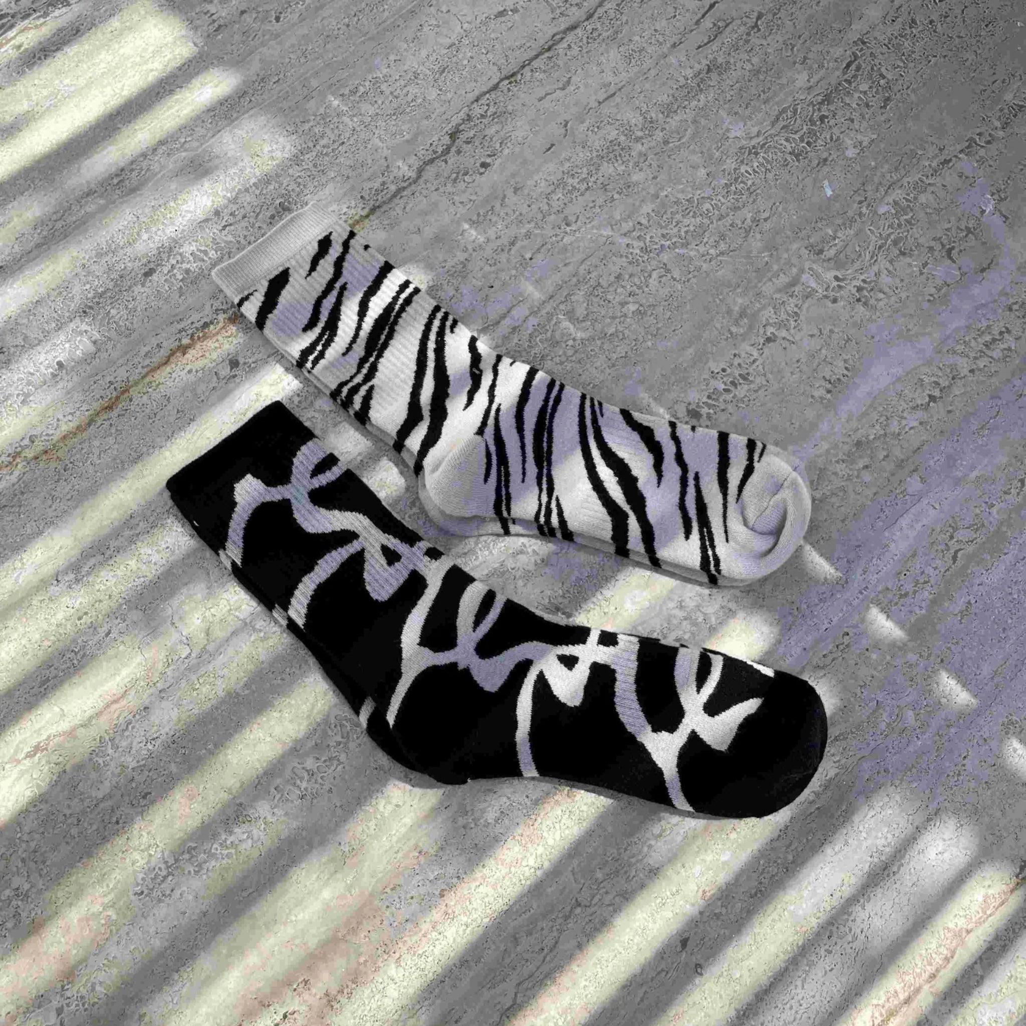 Zwei Socken liegen auf einem Betonboden mit gestreiften Schatten: eine Socke ist weiß mit schwarzen Zebrastreifen, die andere ist schwarz mit einem weißen abstrakten Muster.