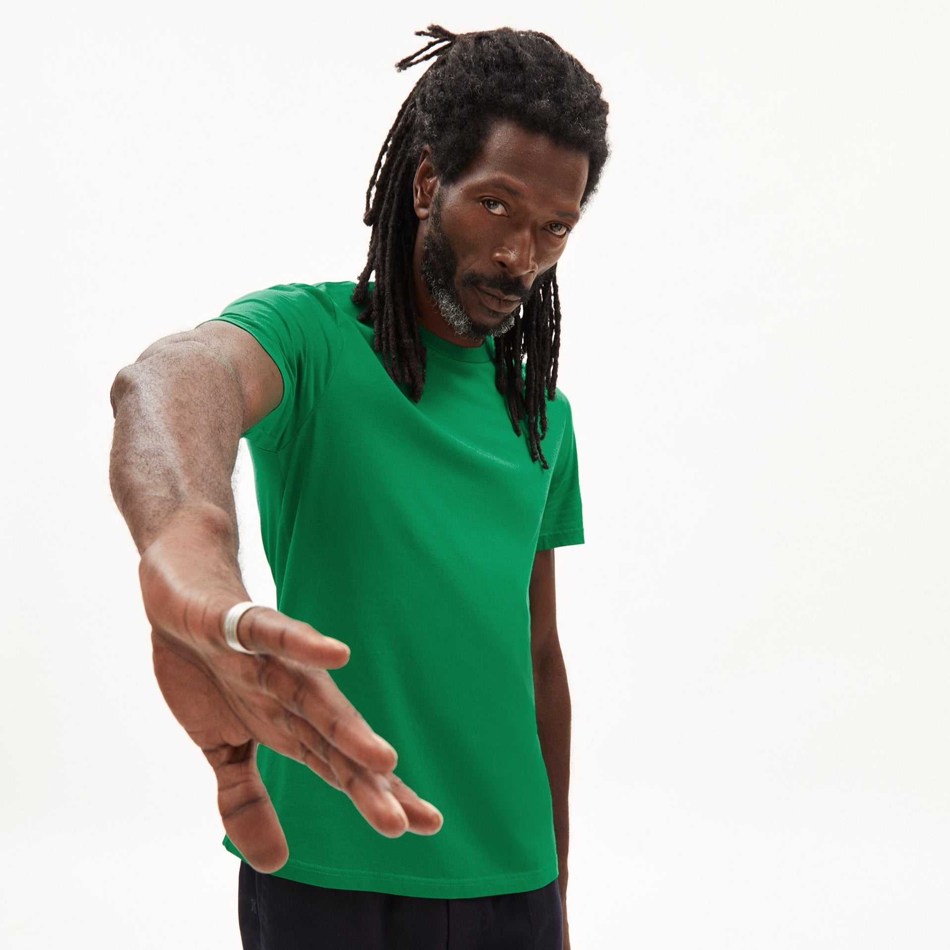Person mit Dreadlocks und grünem T-Shirt streckt einen Arm in die Kamera, vor weißem Hintergrund.