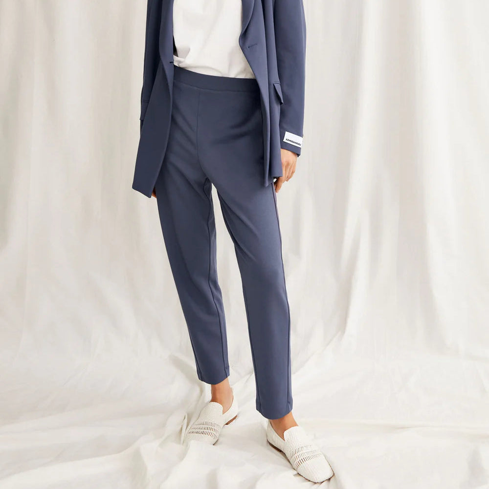 Eine Frau steht vor einem weißen Stoffhintergrund und trägt einen hellvioletten Anzug mit weißem Hemd, weiße Schuhe und eine helle Baseballmütze. Sie nimmt eine selbstbewusste Pose ein und hat eine Hand in ihrer Tasche.