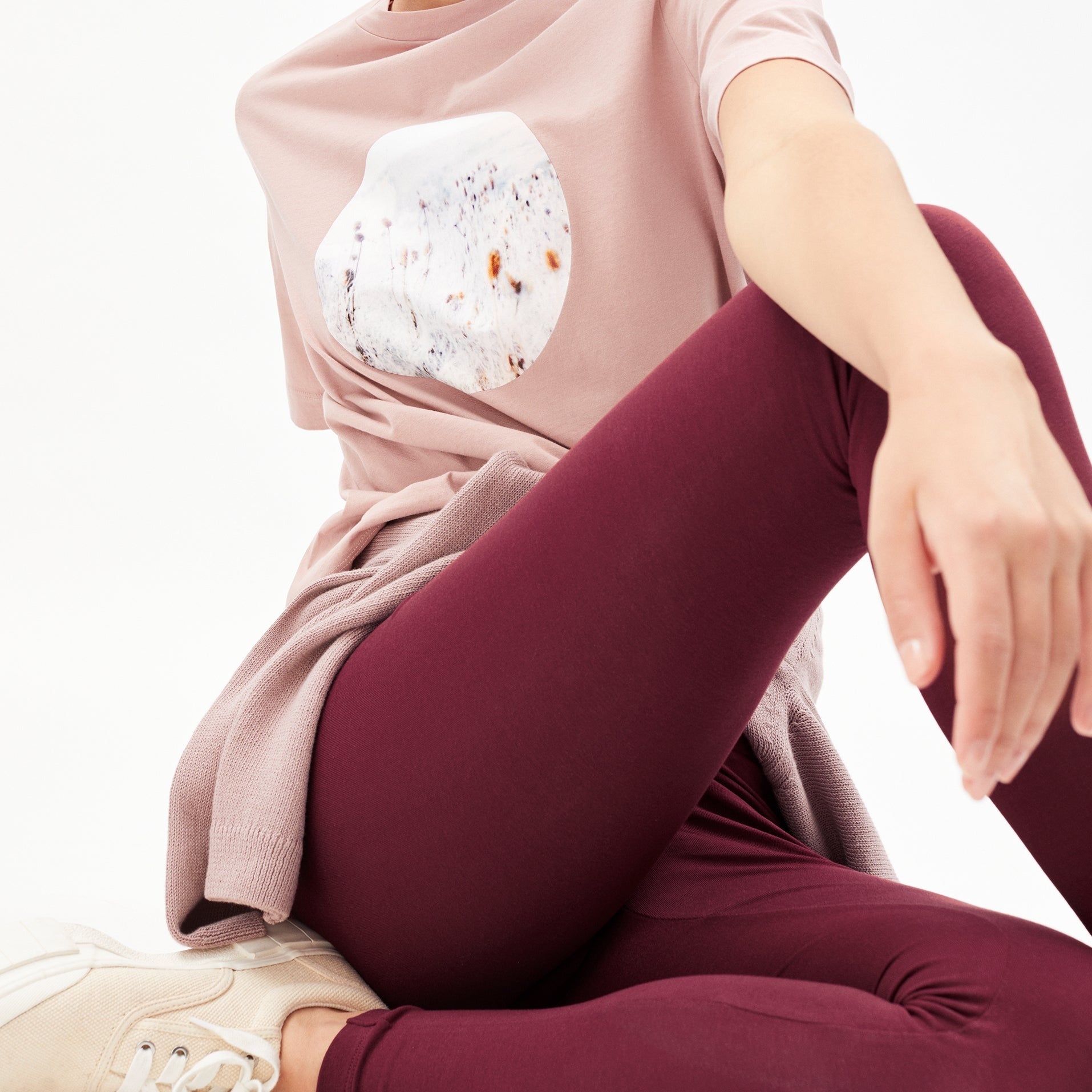 Person in rosa T-Shirt mit abstraktem Muster und bordeauxroter Hose, sitzt mit überkreuzten Beinen auf dem Boden.