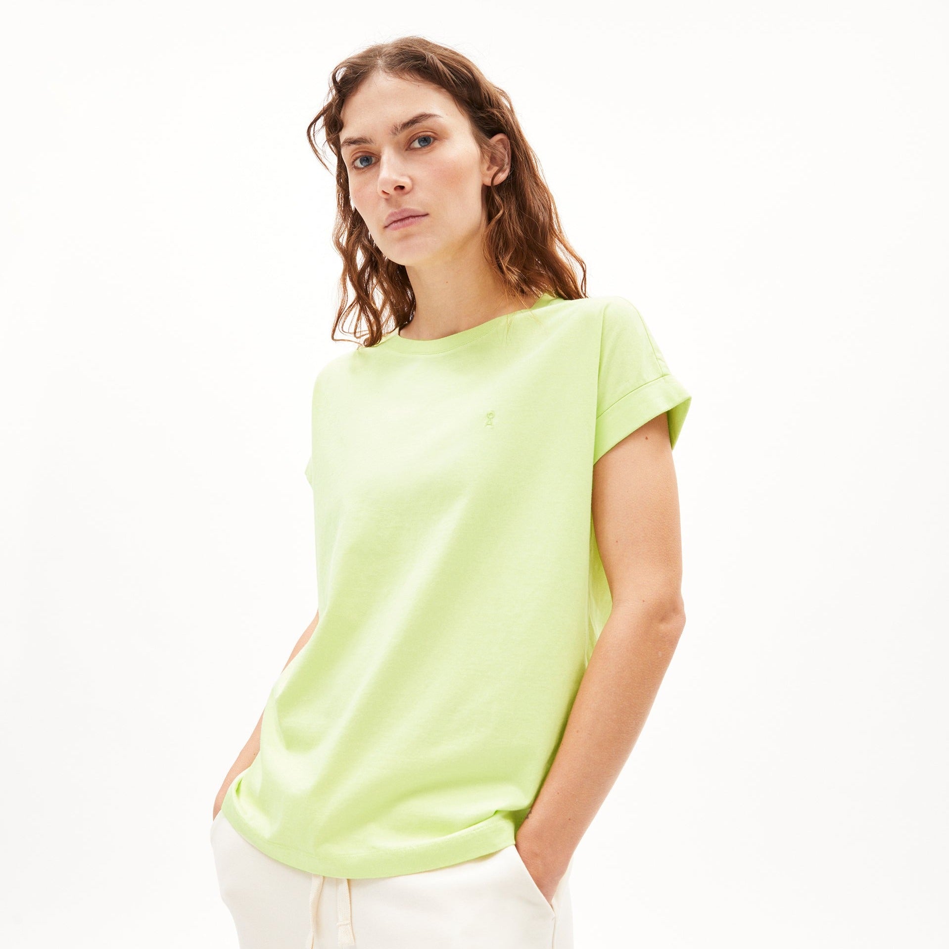 Frau mit grünes T-Shirt und weißer Hose steht vor einem weißen Hintergrund.